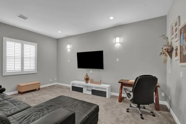 A modern living room with a large flat-screen TV mounted on the wall, a black leather sofa, a wooden desk with a chair, and a small bench.