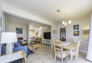 A spacious living area with a sofa, coffee table, and dining table with chairs. There is a television and a chandelier.