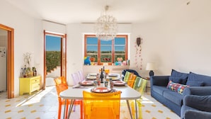 Ein Essbereich mit einem runden Tisch, orangefarbenen Stühlen und einer Kristallleuchter. Im Raum befindet sich ein blaues Sofa und ein gelbes Sideboard.