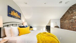 Een slaapkamer met een schuine kap, een bed met wit en geel beddengoed, een bakstenen muur en een schilderij van een gebouw.