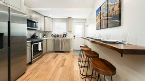 Een moderne keuken met roestvrijstalen apparatuur, houten barstoelen en een schilderij van een brug aan de muur.