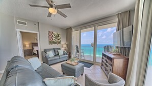 A spacious living room with a large gray sofa, a gray armchair, and a small ottoman. There is a ceiling fan, a TV mounted on the wall, and a view of the ocean through large sliding glass doors.