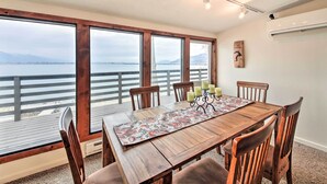 Un comedor con una mesa de madera, seis sillas, un mantel corrido estampado con flores y velas. Amplias ventanas ofrecen vistas al agua y a un puente.