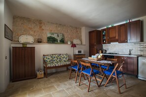 A compact kitchen with a stone wall, wooden cabinets, a dining table with blue chairs, a microwave, and a sink. - 2098 Villa La Mecca by Perle di Puglia (Gagliano del Capo)