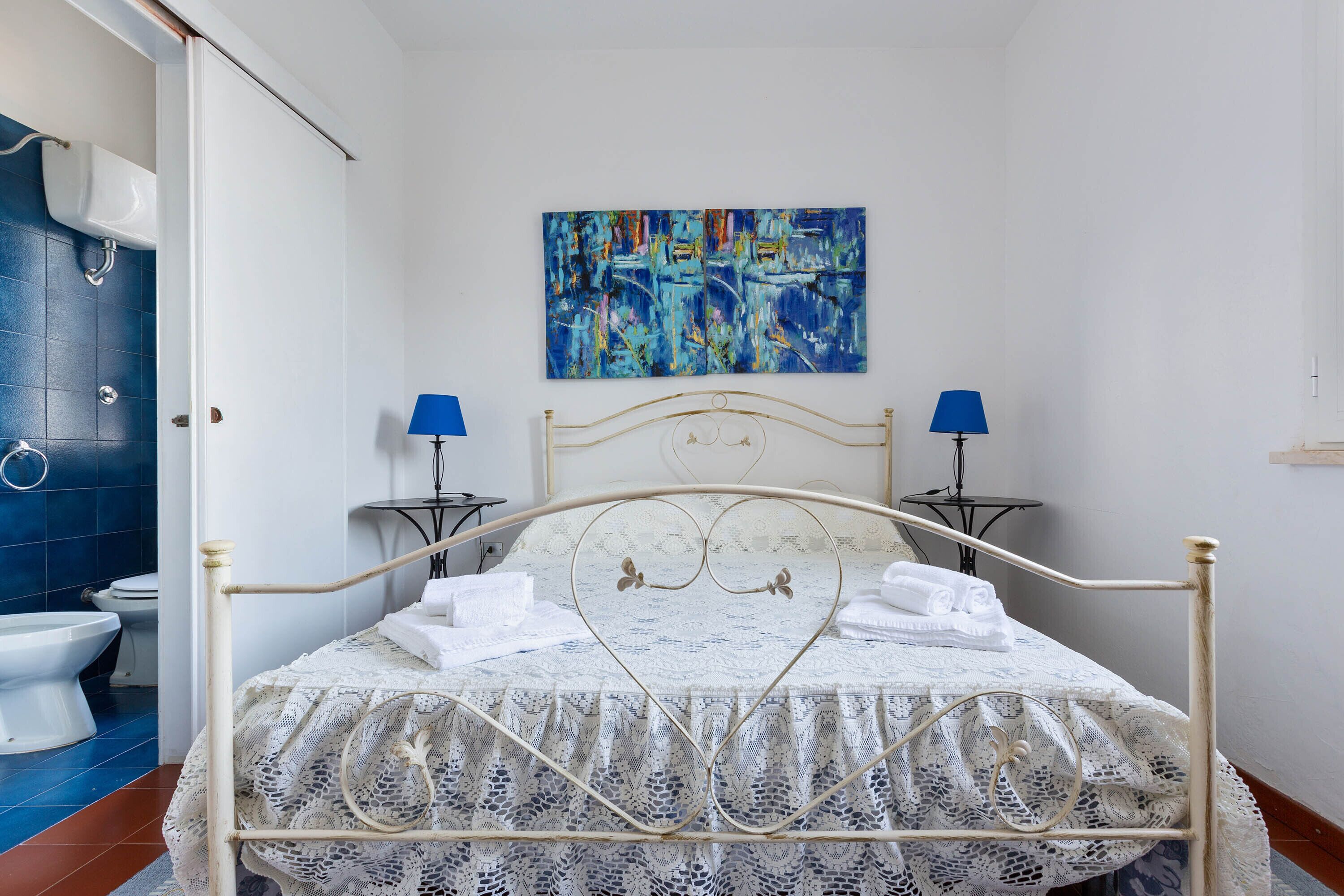 A bedroom with a white metal bed frame, a lace bedspread, two bedside lamps, and a painting on the wall.