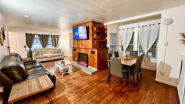 A living room with a wooden floor, a leather sofa, a TV mounted on a wooden cabinet, a fireplace, and a dining area with a table and chairs.