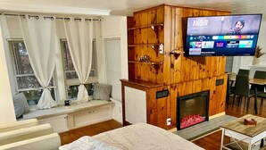 A bedroom with a large bed, a wooden wall unit with a TV, a fireplace, and a seating area by the window.