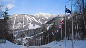 A snowy mountain road with flags, a sign reading "Welcome Whiteface Olympic Mountain 3430 Vertical", and a speed limit sign of 20.
