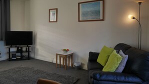 A living room with a black leather sofa, a small wooden side table with a bowl of fruit, a black TV on a stand, and two framed pictures on the wall. - Cliffwalk Cottage (Portland)