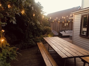 A wooden deck with a picnic table and a wicker chair, surrounded by greenery and string lights.