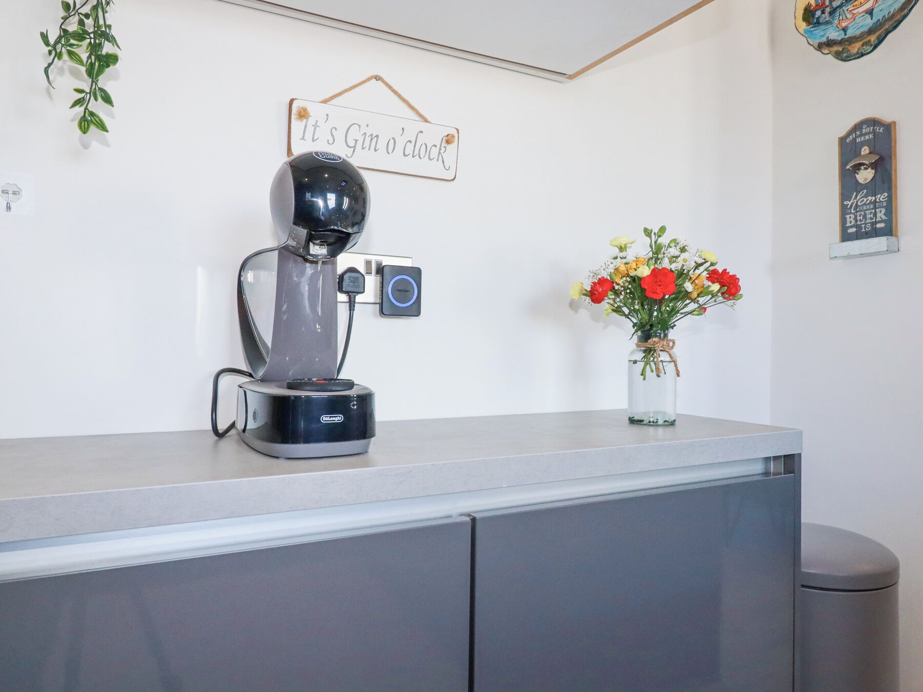 A coffee machine on a counter, a sign that says "It's Gin o'clock", a small vase with flowers, and a wall decoration.