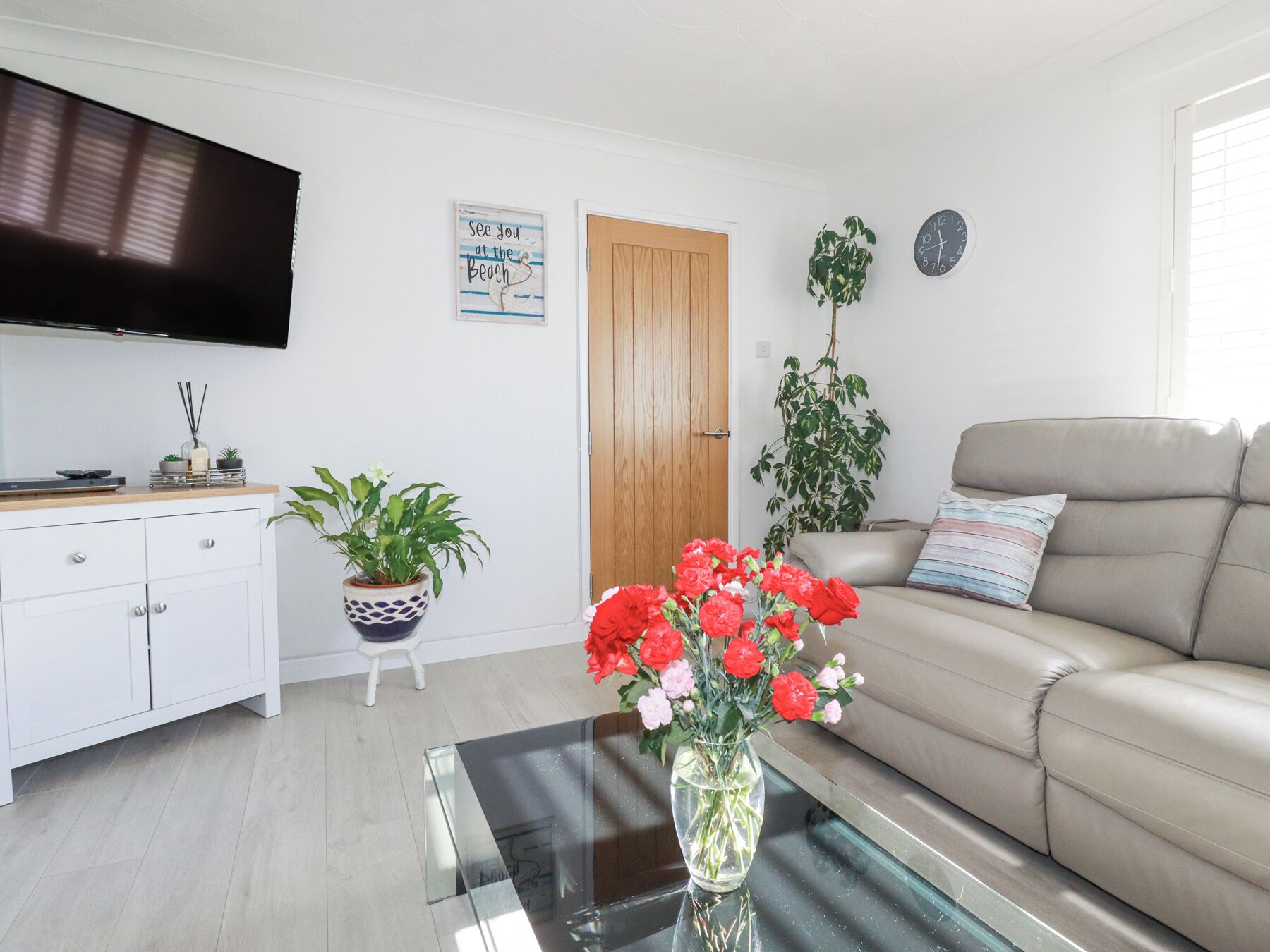 Un salon moderne avec un téléviseur à écran plat, une porte en bois, un canapé beige et une table basse en verre sur laquelle est posé un vase de fleurs.