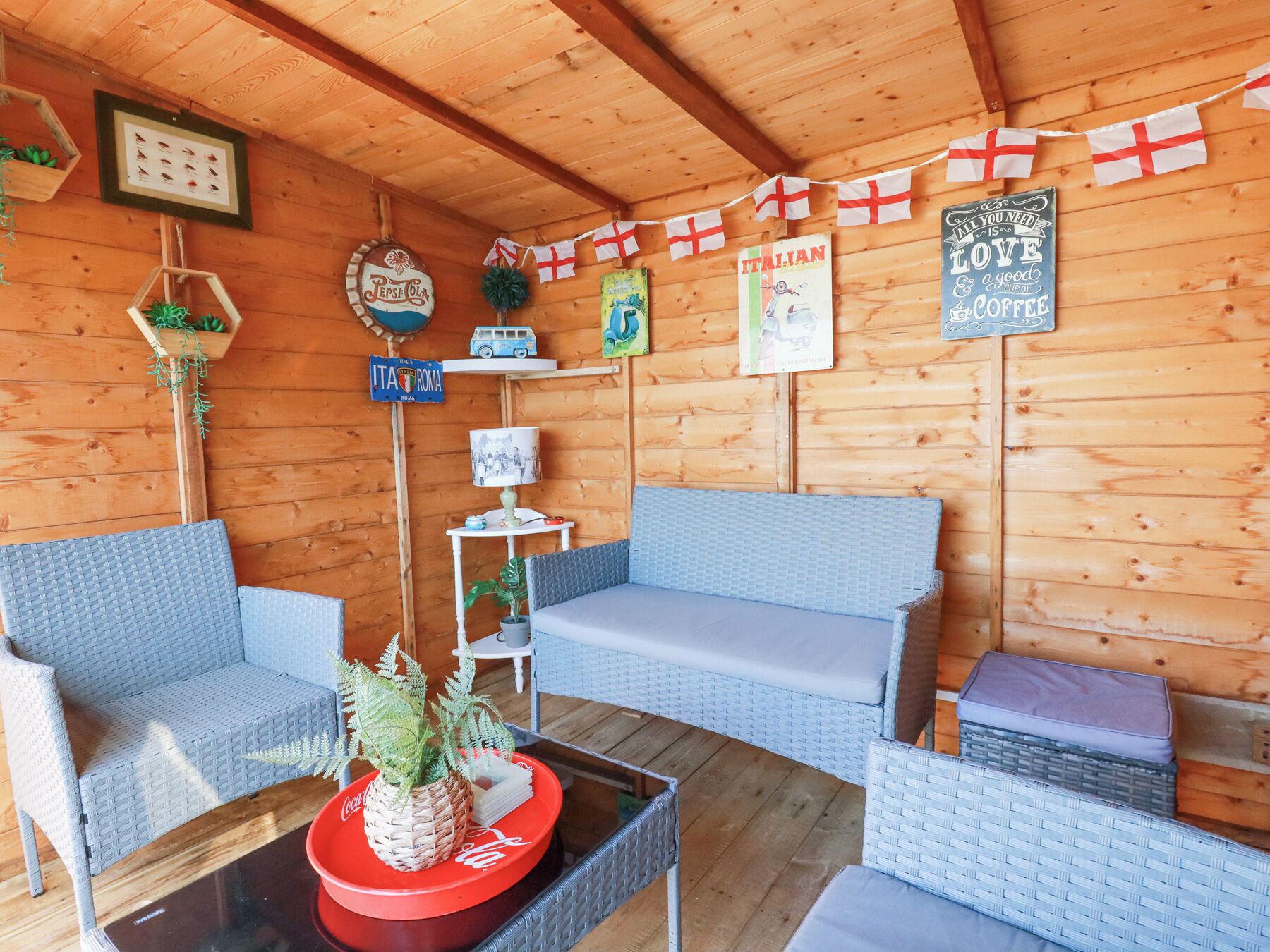 Una cabaña de madera acogedora con mobiliario de mimbre, una bandeja roja de Coca-Cola y varios objetos decorativos, incluyendo un letrero de Pepsi Cola, una bandera italiana y un cartel que dice 'Todo lo que necesitas es amor y una taza de café'.
