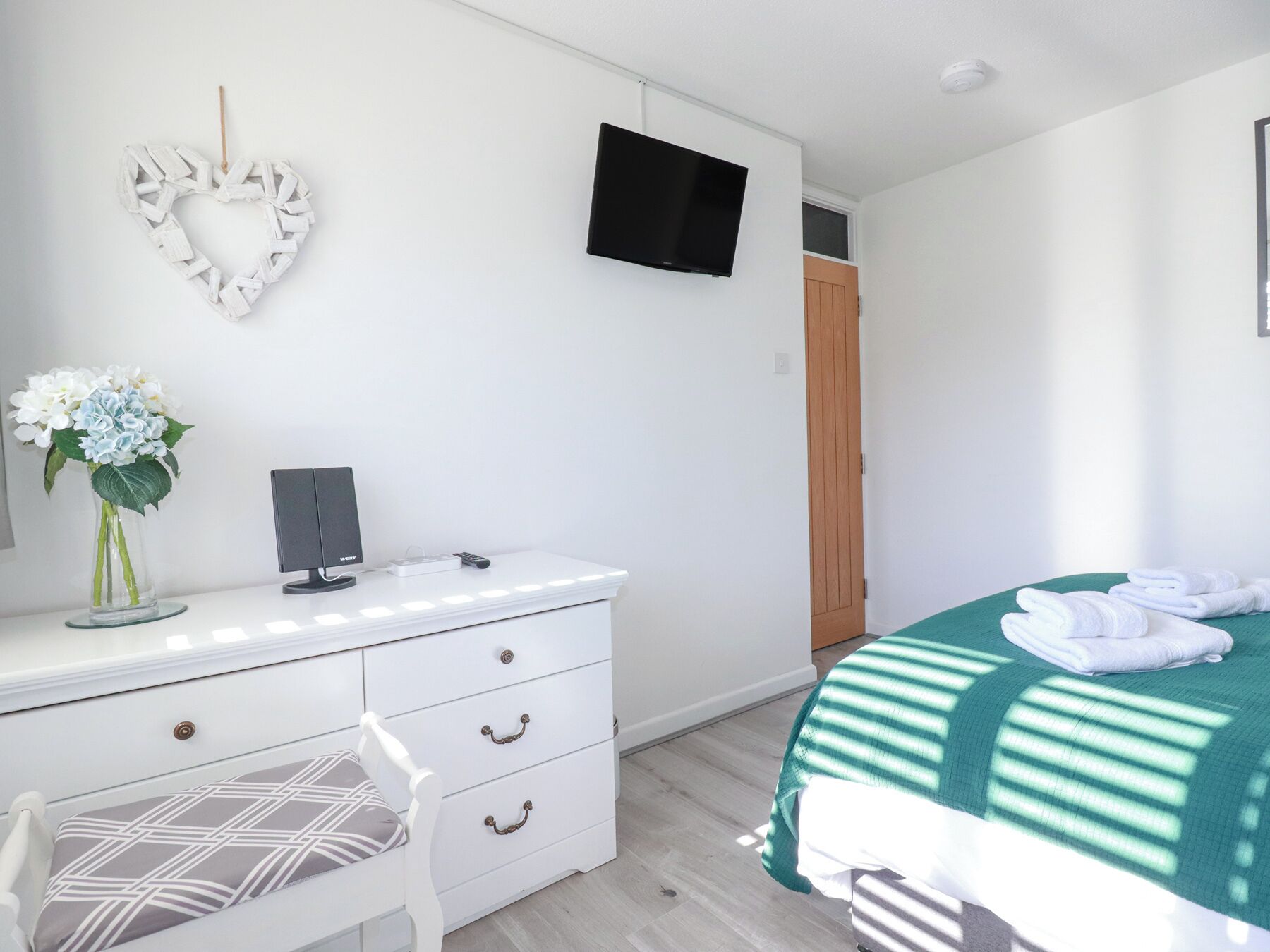 Un dormitorio con un aparador blanco, un televisor montado en la pared, una decoración con forma de corazón, una cama con una manta verde y una puerta de madera.