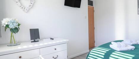 Un dormitorio con un aparador blanco, un televisor montado en la pared, una decoración con forma de corazón, una cama con una manta verde y una puerta de madera.