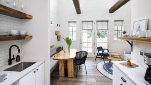 A modern kitchen with white cabinetry, a wooden dining table, and a black chair. There is a window with a view of greenery outside. - Elevated, Boutique Space on the Swamp Rabbit Trail (Travelers Rest)