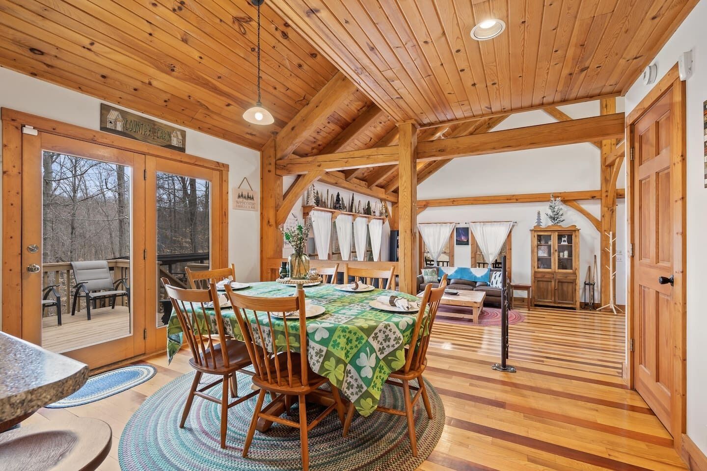 A wooden cabin interior with a dining area, a living room, and a view through a door to an outdoor deck.