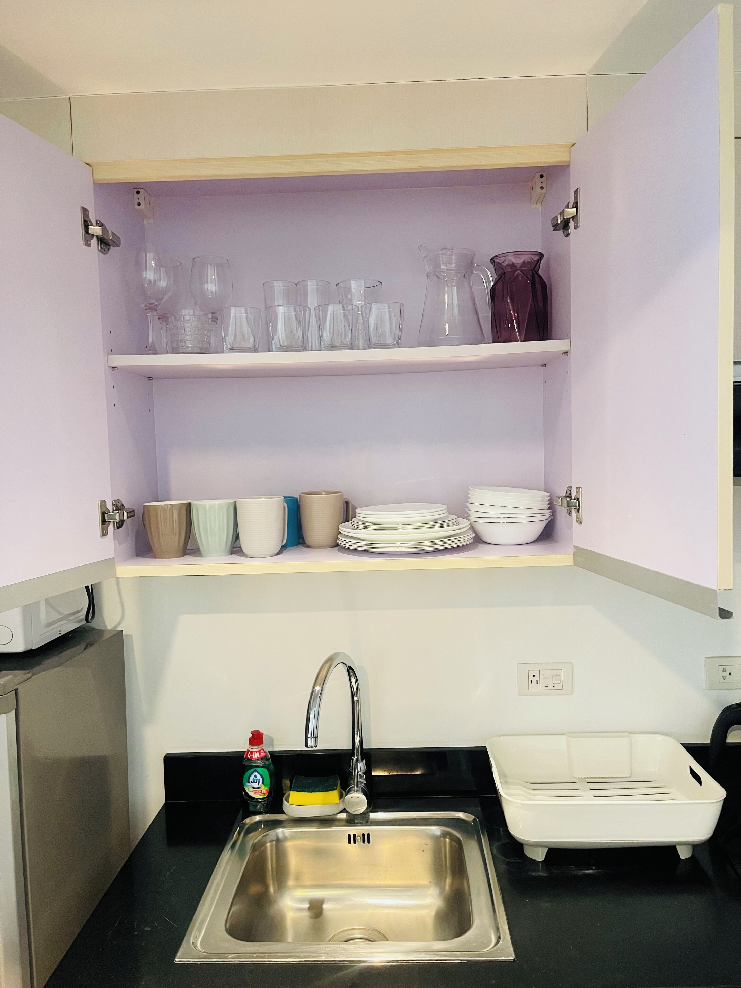 Una cocina con un lavamanos de acero inoxidable, un escurreplatos y estantes superiores con cristalería y vajilla.