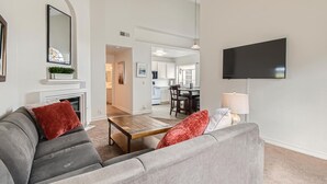 A living room with a grey sofa, a wooden coffee table, and a fireplace. A television is mounted on the wall, and a kitchen is visible in the background. - 1 Block to Pacific Beach/FOOD/Bars, Updated, 2-Car Garage w/fee, Yard, Sleeps 14 (San Diego)