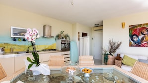 A living room with a glass coffee table, wicker furniture, a painting, and a kitchen in the background. - 2539 Casa di Isa by Barbarhouse (Castrignano del Capo)