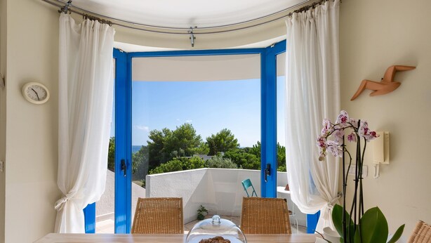 A room with a glass table, two wicker chairs, a glass cake stand, a clock, and a view of greenery outside. - 2539 Casa di Isa by Barbarhouse (Castrignano del Capo)