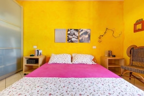 A room with a bed, two nightstands, a wicker chair, and a wall with three artworks.