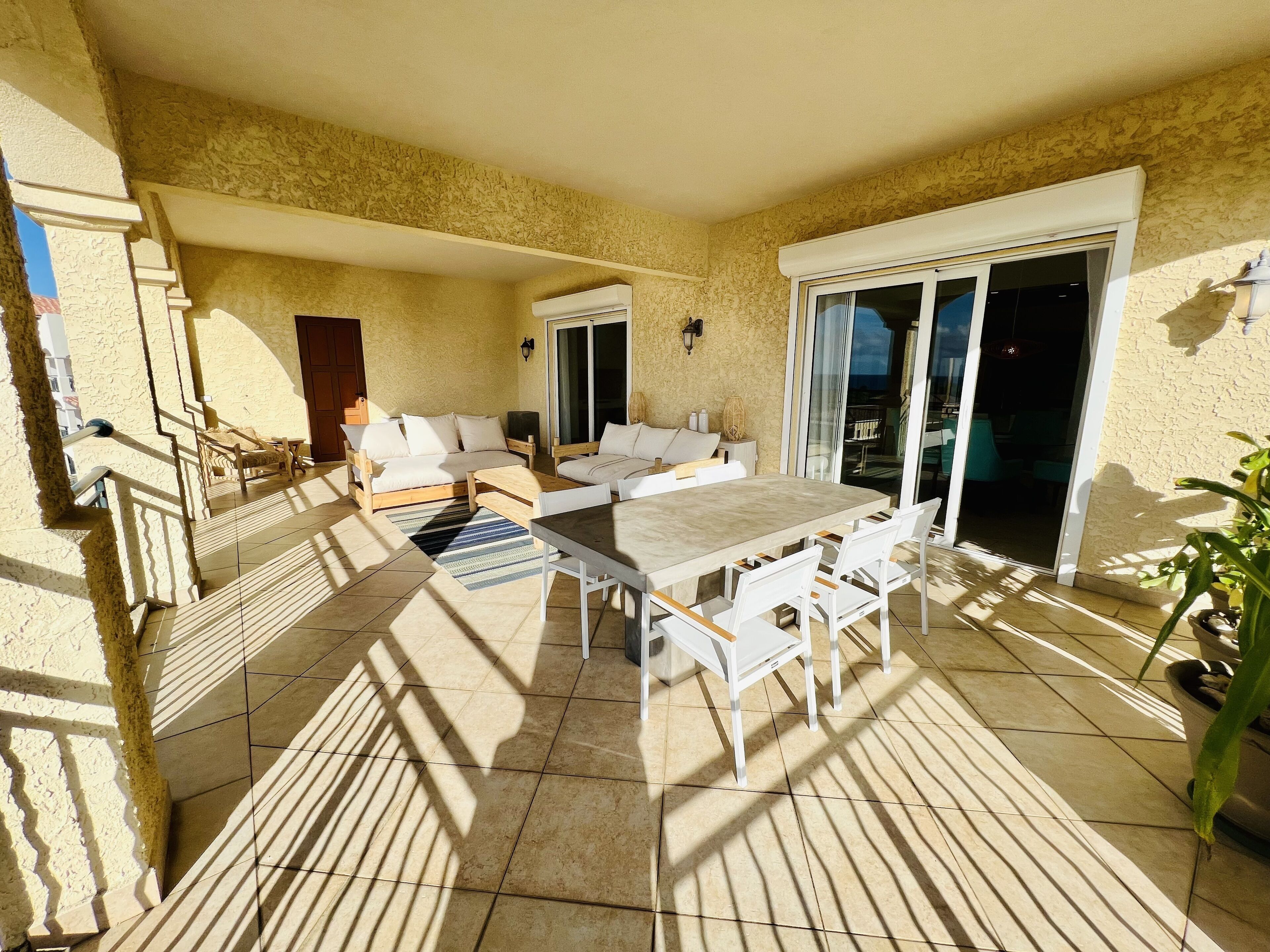 A sunlit patio with a dining table and chairs, a sofa, and a sliding glass door leading to an outdoor area.