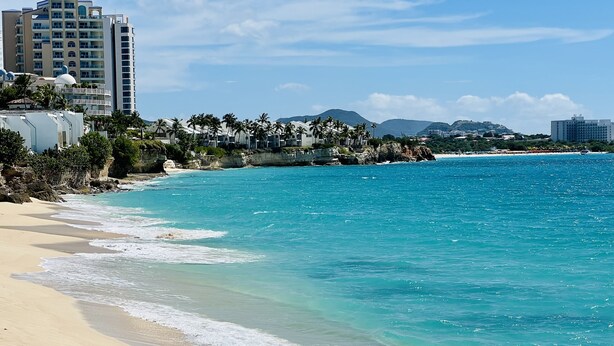 Una playa con agua turquesa cristalina, orilla de arena y edificios al fondo.