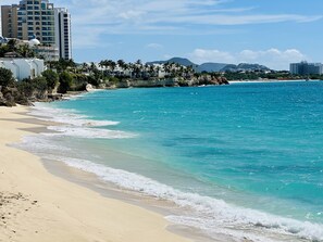 Una playa con agua turquesa cristalina, orilla de arena y edificios al fondo.