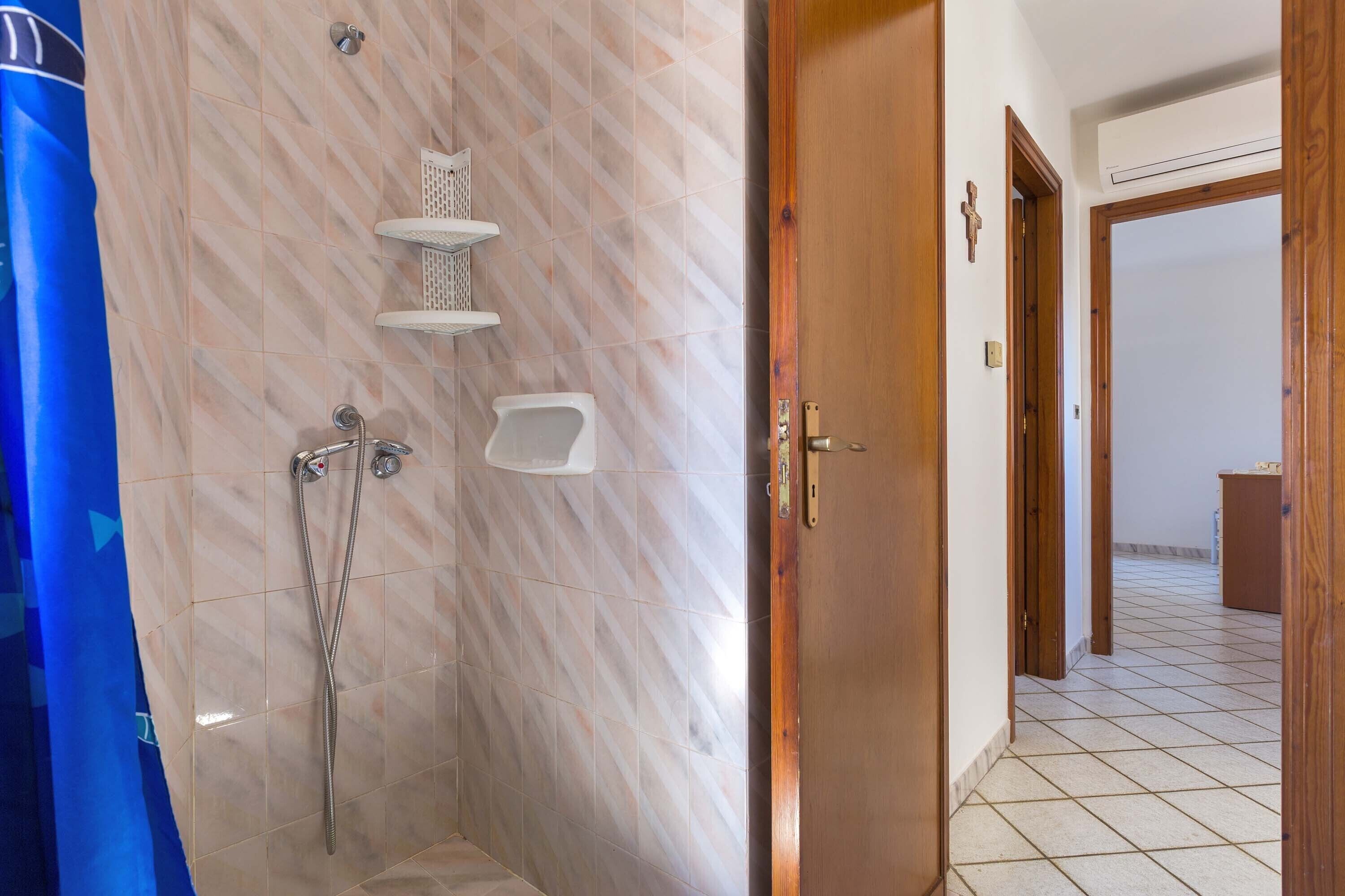 A bathroom with a shower, a toilet, and a sink. There is a wooden door and a tiled floor.