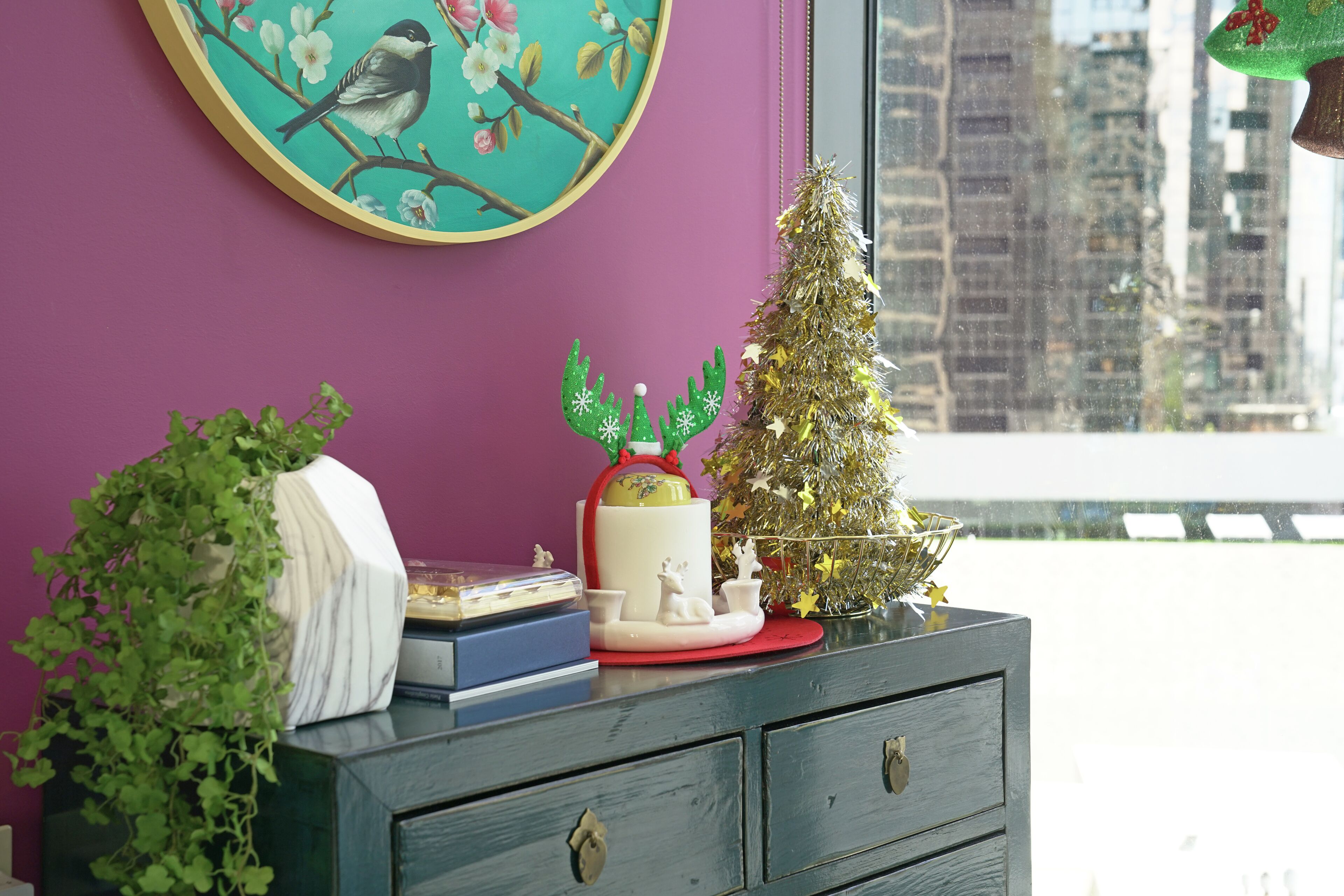 A room with a purple wall, a window with a city view, a small Christmas tree, and a decorative plate with a reindeer.
