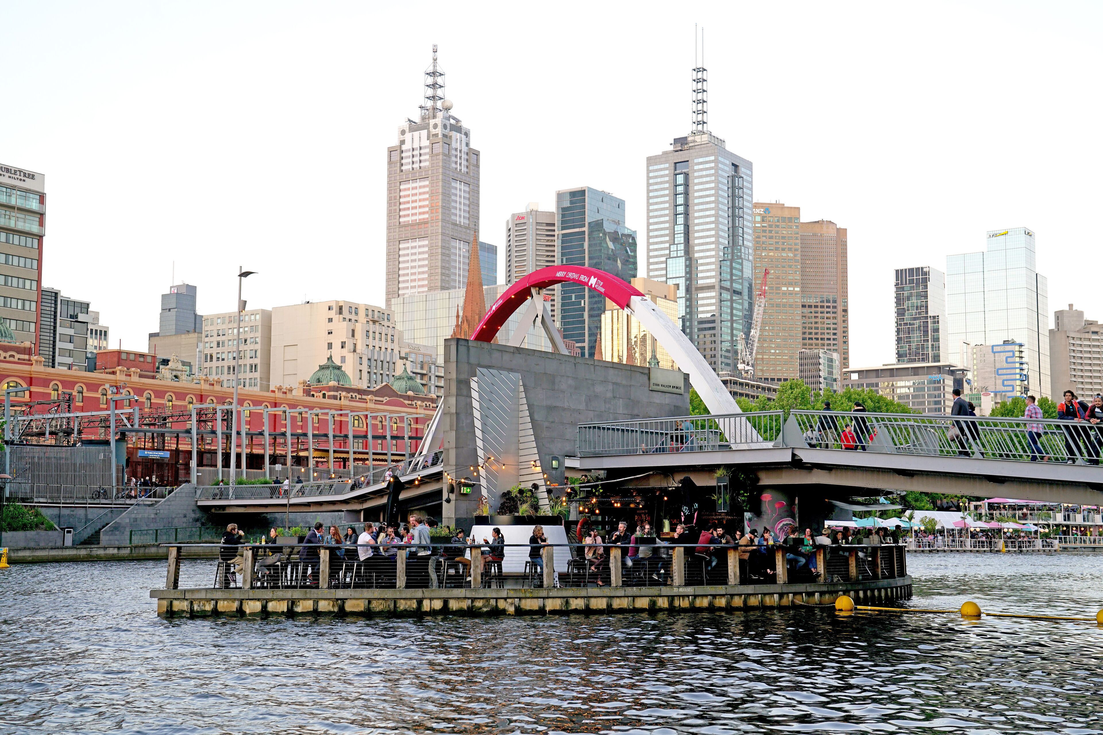 A riverside restaurant with outdoor seating, a bridge, and a city skyline in the background.
