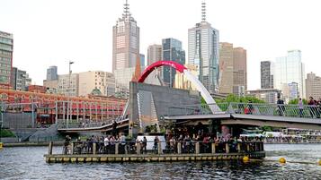 A riverside restaurant with outdoor seating, a bridge, and a city skyline in the background.