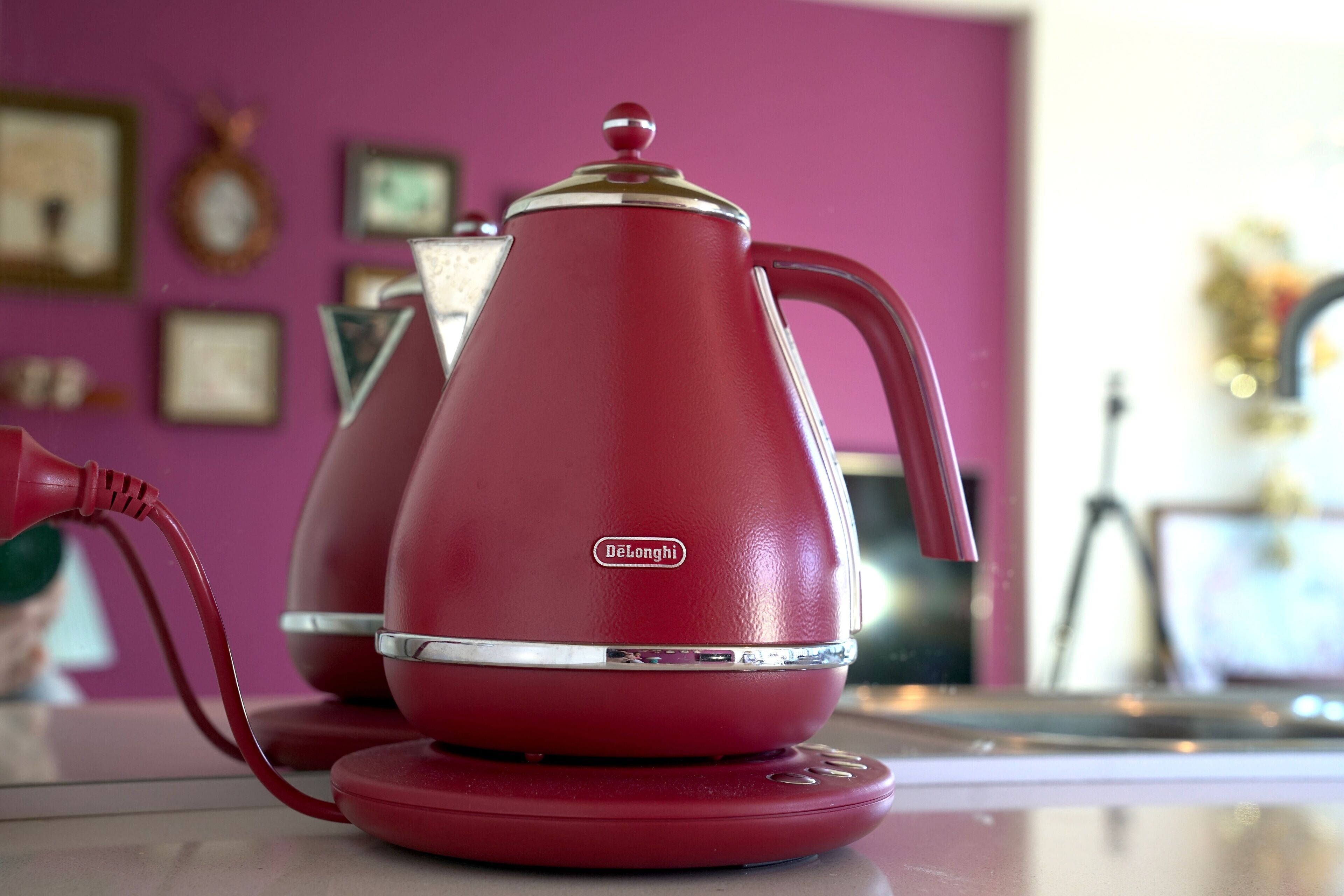 A red DeLonghi coffee maker on a kitchen counter with a pink wall in the background.