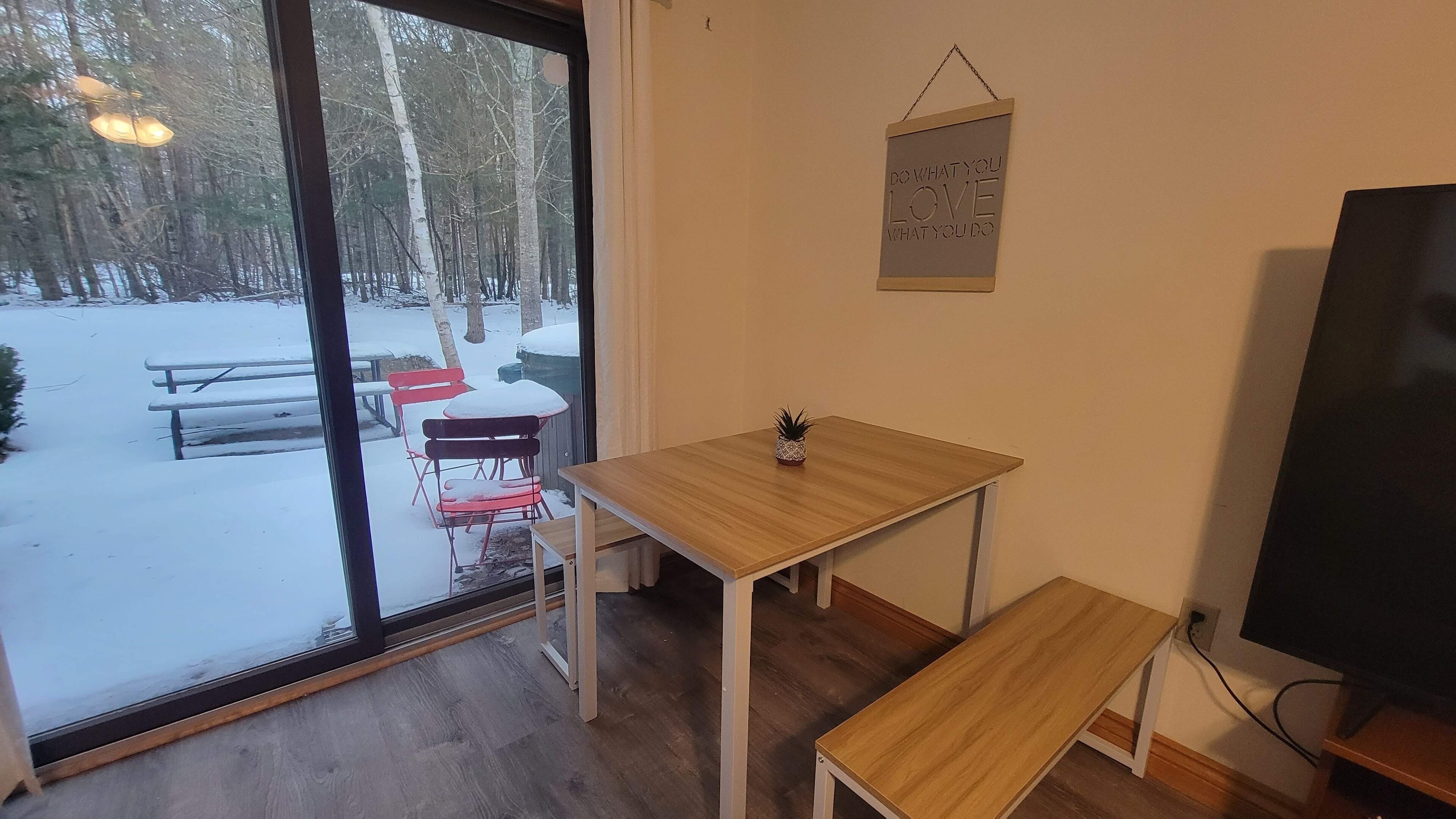A room with a wooden table and bench, a small potted plant, a TV, and a window with a view of a snowy outdoor area with benches and trees.