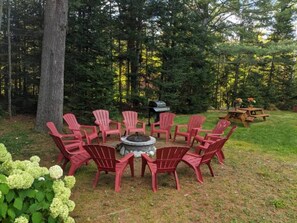 A circular seating arrangement with red chairs around a central fire pit, set in a grassy area with trees in the background. - Charming Guest House & King Suite - Twin Mountain River Retreat (Carroll)