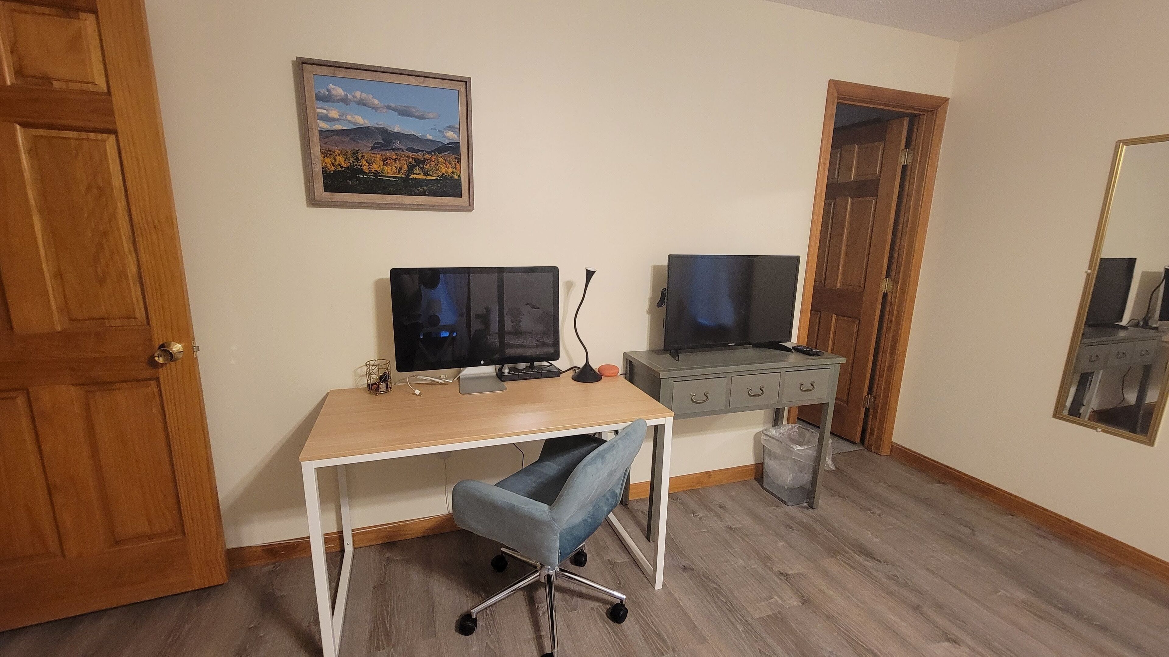 Ett rum med ett skrivbord i trä, två datorskärmar, en stol, en liten papperskorg och en dörr.