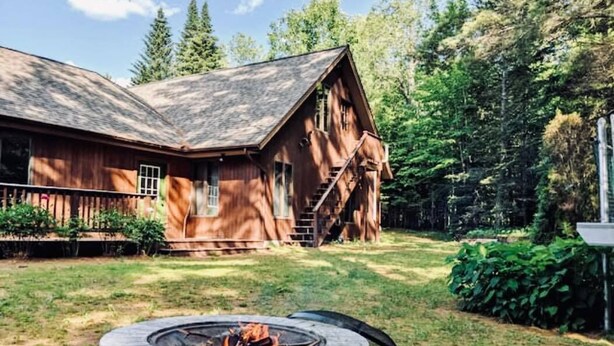 Une cabane rustique avec un foyer en pierre en premier plan, entourée d’un jardin bien entretenu et de grands arbres.