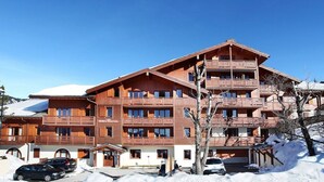 A multi-story wooden building with balconies, a ski slope in the background, and snow on the ground.