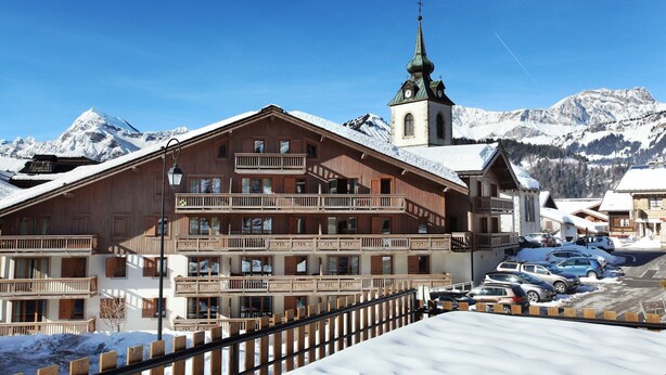 A wooden building with multiple balconies and a church with a spire, surrounded by snow-covered mountains.