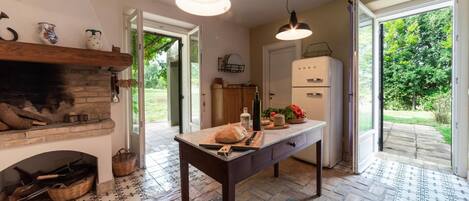 Una cocina con chimenea de ladrillo, una mesa de madera con pan y un cuchillo, un refrigerador y vistas de un jardín a través de la puerta.