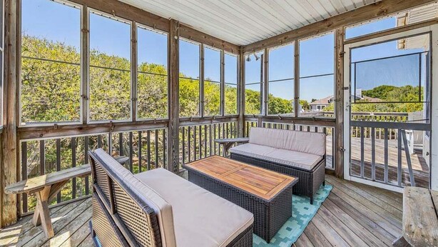 A covered outdoor deck with wicker furniture, a wooden table, and a view of greenery and houses. - A Peace of Quiet | 1,025 ft. to the Beach | Private Pool (Duck)