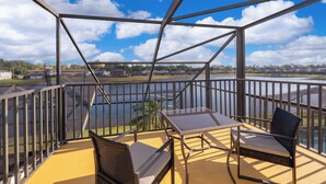 Uma varanda com guarda-corrimão de metal, duas cadeiras, uma mesa pequena e vista para um lago e casas ao fundo.