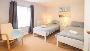 A bedroom with two beds, a chair, a radiator, and a window with curtains. - Pebble Reef (Portland)