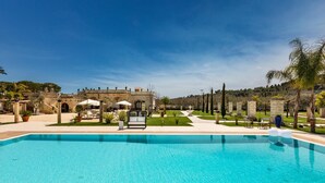 Ein Swimmingpool mit klarem, blauem Wasser, umgeben von einem angelegten Garten, einem Gebäude mit Bögen und einer Palme.