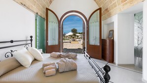Ein Schlafzimmer mit Bett, Kissen, Handtüchern und Blick durch einen Bogen zur Außenanlage.