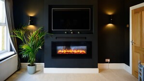 Una sala de estar moderna con una chimenea negra, un televisor de pantalla plana montado encima y una planta en maceta a la izquierda.