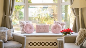 Un salón acogedor con un sofá, un sillón y una mesita. Se ve un rincón con cojines junto a la ventana, y una lámpara colgante arriba.