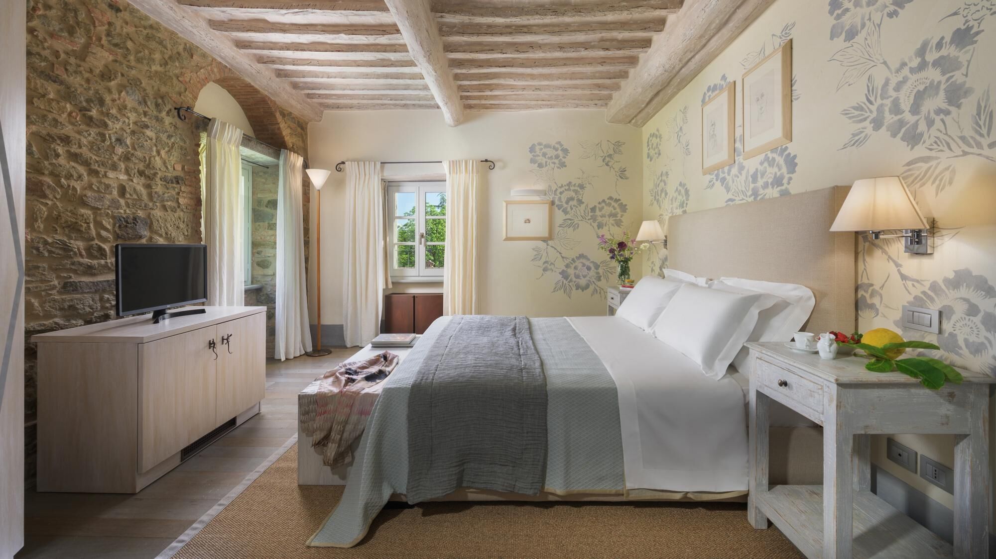A bedroom with a stone wall, a bed with white linens, a TV on a wooden cabinet, and a window with curtains.