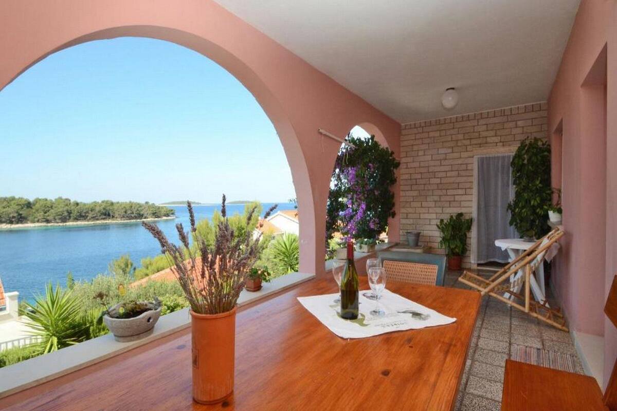 A balcony with a view of the sea, a wooden table with a bottle and glasses, and potted plants.